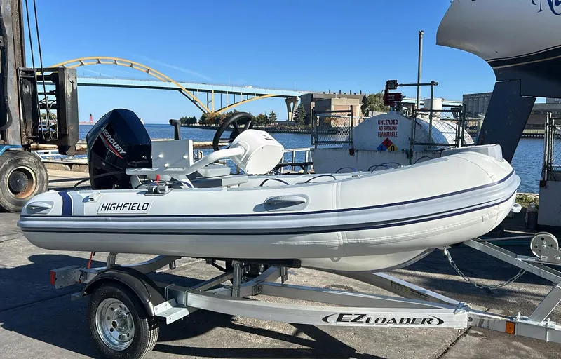The Image of 2025 Highfield CL340 inflatable boat on trailer by waterfront, featuring Mercury outboard motor. - 0