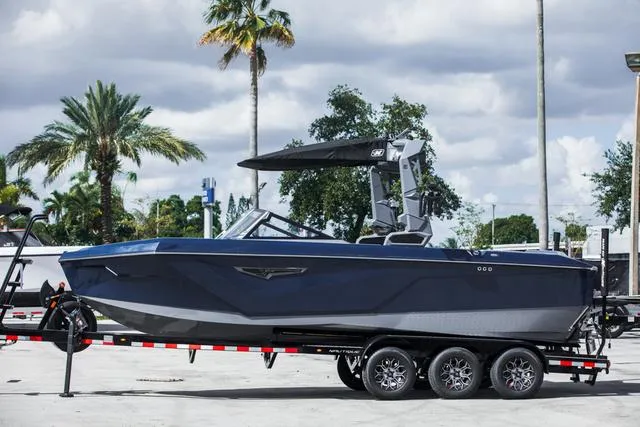 Slide: The Image of 2024 Nautique S23 boat on trailer, parked outdoors with palm trees in background. - 2