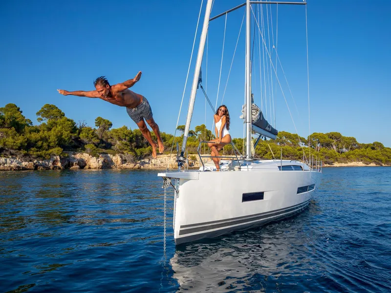 Slide: The Image of Man diving off 2025 Hanse 410 yacht, woman seated, scenic coastal backdrop. - 13
