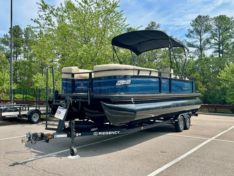The Image of 2025 Regency 250 DL3 pontoon boat on trailer in parking lot, surrounded by trees. - 0