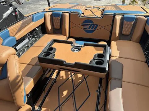 Slide: The Image of 2025 Malibu M230 boat interior with tan and blue seating, featuring a central table and cup holders. - 23
