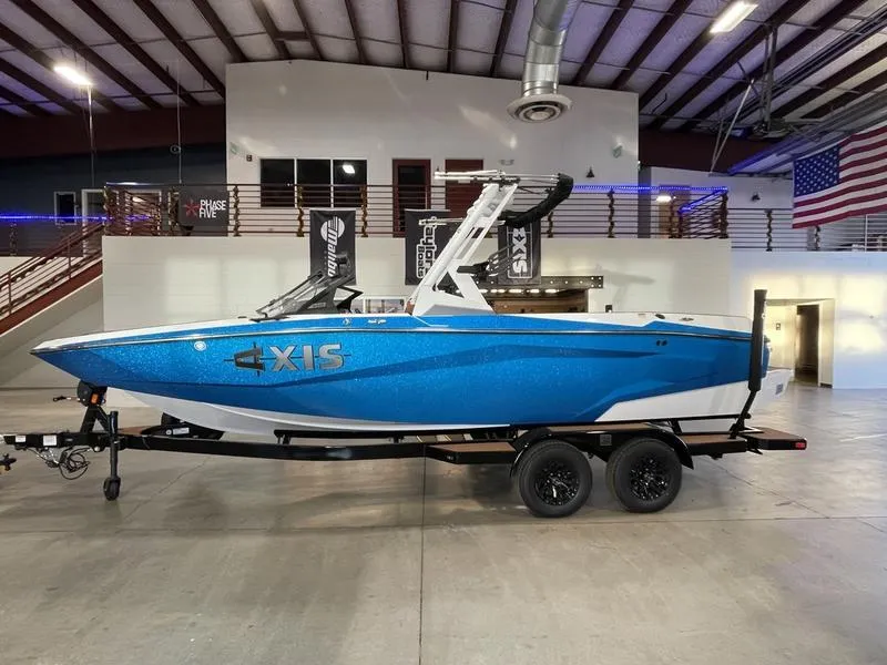 Slide: The Image of 2025 Axis A225 boat in showroom, blue exterior, on trailer, American flag in background. - 1