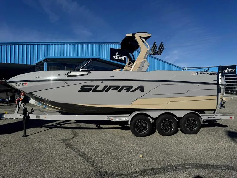 Slide: The Image of 2025 Supra SE boat on trailer, parked outdoors near blue building. - 0
