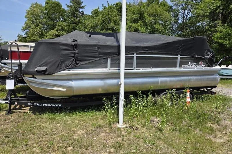 The Image of 2025 Sun Tracker PB 22 RF pontoon boat covered, parked on trailer, surrounded by greenery. - 2