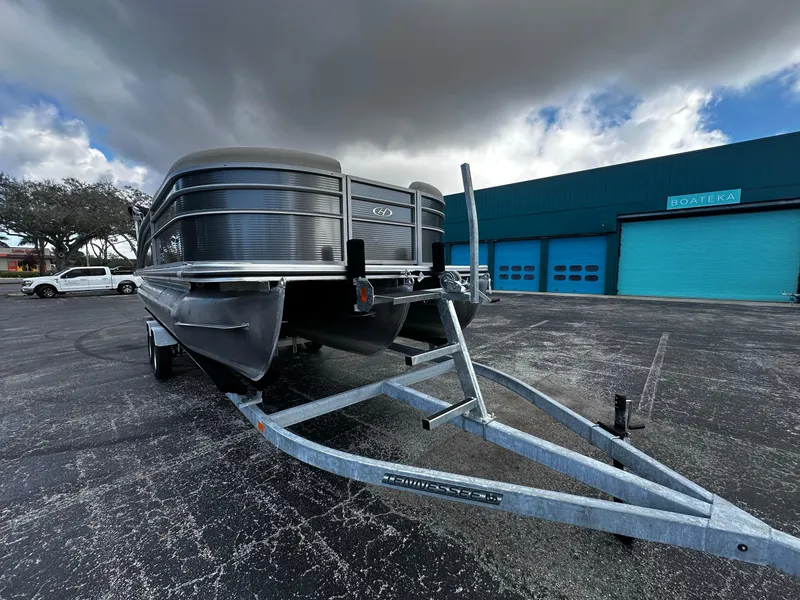 Slide: The Image of 2023 Harris Cruiser 230 pontoon boat on trailer, parked outside a blue building. - 3