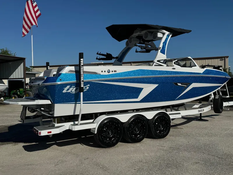 Slide: The Image of 2025 Tigé 24RZX boat on trailer, blue and white design, parked outdoors. - 2