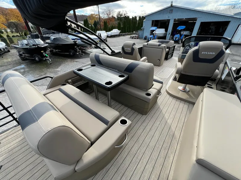 Slide: The Image of 2025 Sylvan L3 DLZ Tritoon interior with beige seating and table, parked near a garage. - 9