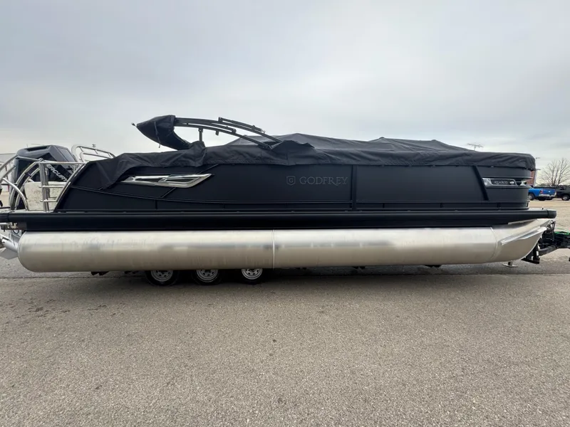 Slide: The Image of 2025 Godfrey AQUAPATIO 255SFL pontoon boat with black cover, parked on pavement. - 3