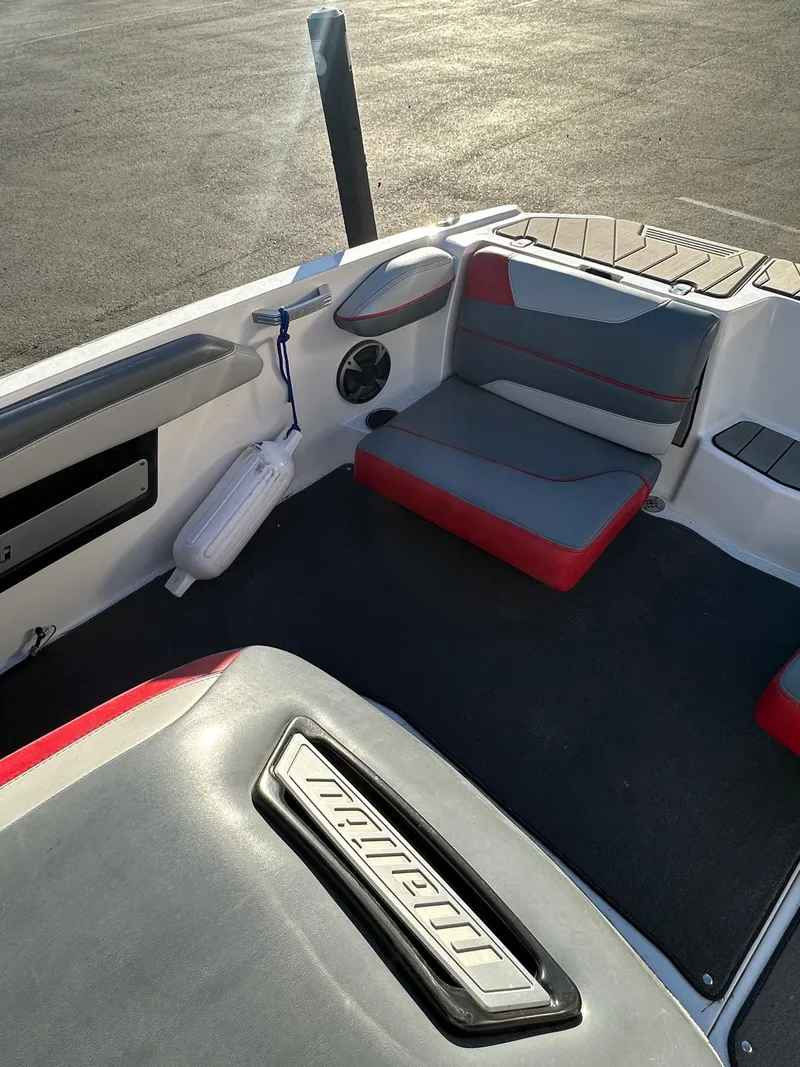 Slide: The Image of 2019 Malibu TXi Mo boat interior with gray and red seating, docked on asphalt. - 12