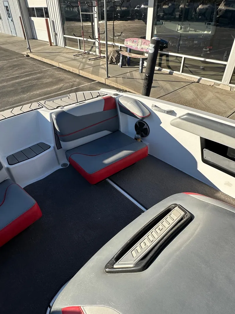 Slide: The Image of 2019 Malibu TXi Mo boat interior with gray and red seating, docked near a building. - 13