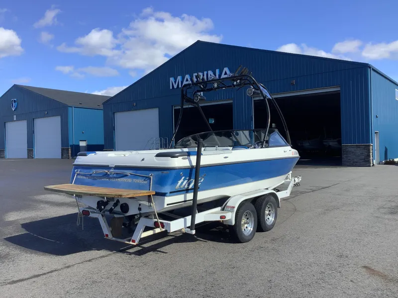 Slide: The Image of 2002 Tigé 22i boat on trailer outside marina, blue and white design. - 7