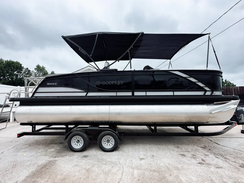 Slide: The Image of 2025 Godfrey Monaco 23' Dual Flip Lounge Tritoon on trailer, black canopy, parked outdoors. - 17