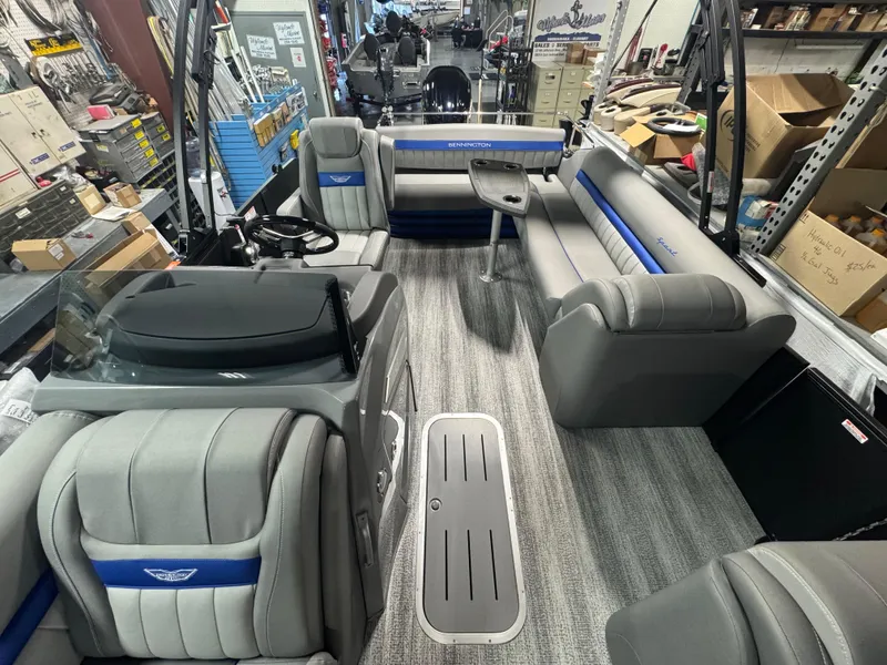 Slide: The Image of This is an image of the interior of a pontoon boat. The boat features a spacious seating area with cushioned seats in a grey and blue color scheme. The floor is covered with a grey carpet, and there is a small table in the center. The boat appears to be w - 6
