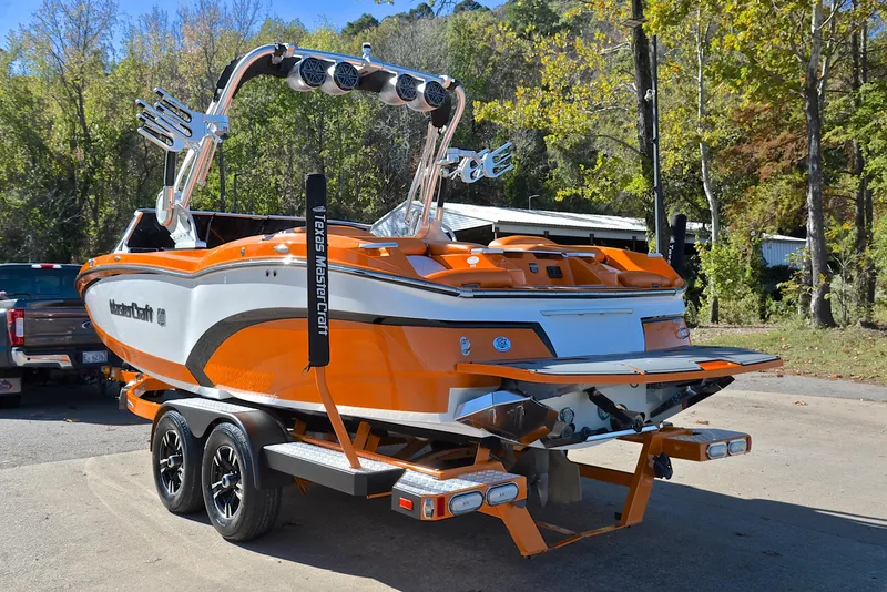 Slide: The Image of 2015 MasterCraft X23 boat in vibrant orange, parked on a trailer, surrounded by trees. - 12