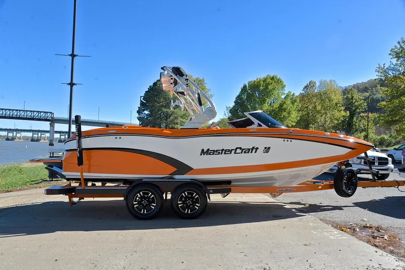 Slide: The Image of 2015 MasterCraft X23 boat on trailer by riverside, vibrant orange and white design. - 15
