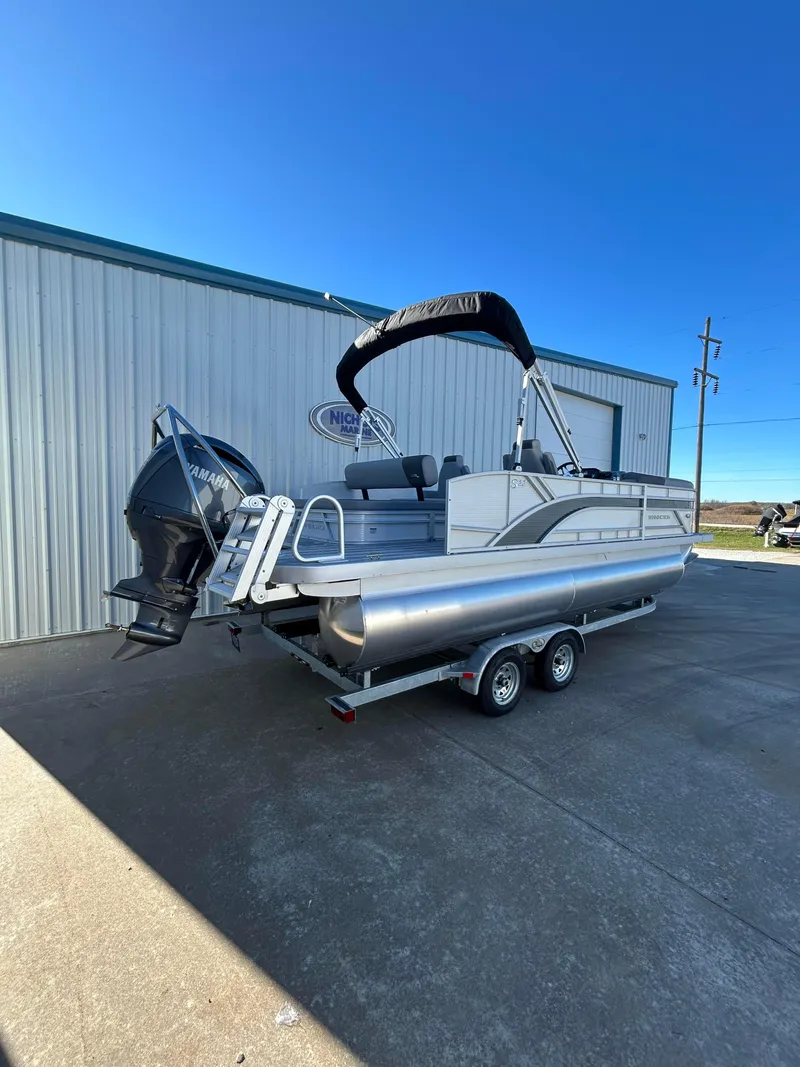 Slide: The Image of 2025 Bennington S 22 pontoon boat on trailer, parked outside a metal building. - 2