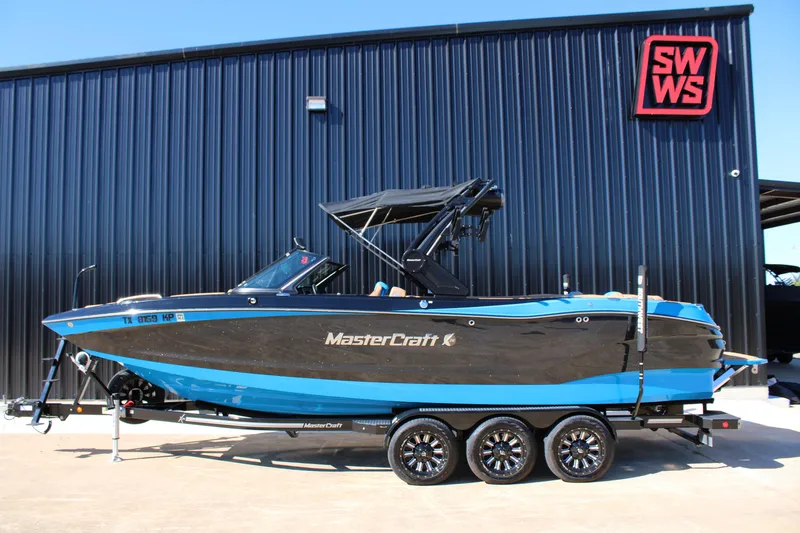 The Image of 2023 MasterCraft X26 boat in blue and black, parked on a trailer outside SWWS building. - 1