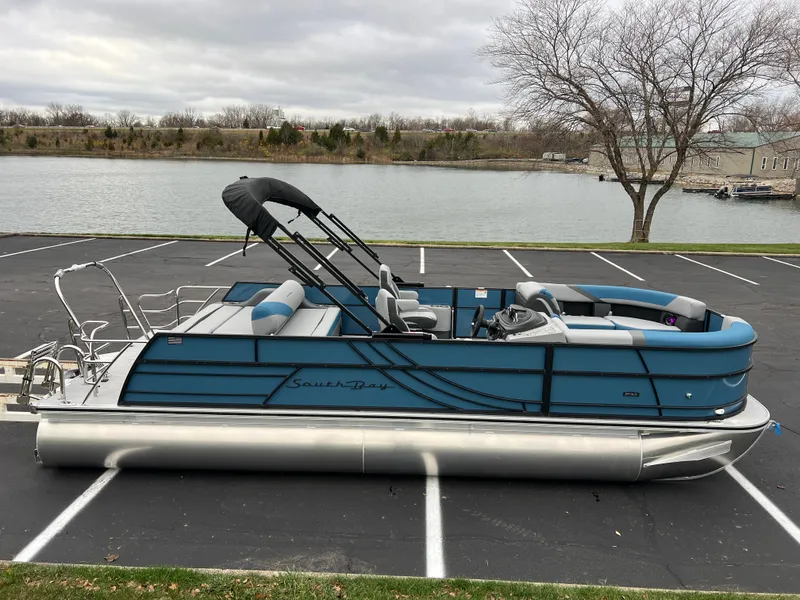 Slide: The Image of 2025 South Bay S224UL pontoon boat parked near a lake on a cloudy day. - 0