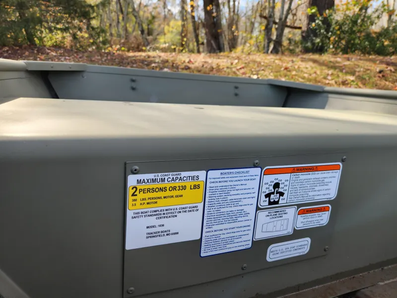 Slide: The Image of 2025 Tracker Grizzly 1036 Utility boat with capacity and safety labels in a wooded area. - 3