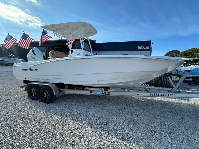 Slide: The Image of 2025 NauticStar 231 Hybrid boat on trailer, flags in background, sunny day. - 1