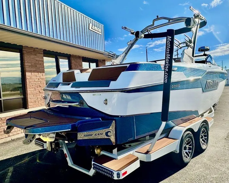 Slide: The Image of 2023 Malibu M220 boat on trailer, parked outside a building under a clear blue sky. - 2