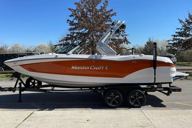 The Image of 2024 MasterCraft X22 boat on trailer, orange and white design, parked outdoors. - 0
