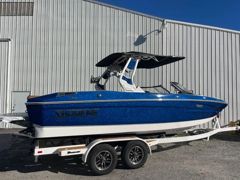 Slide: The Image of 2024 Supreme S220 boat in blue on a trailer, parked outside a metal building. - 13