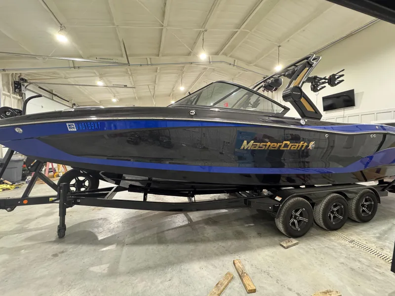 The Image of 2024 MasterCraft X26 boat in a garage, featuring a sleek black and blue design. - 0