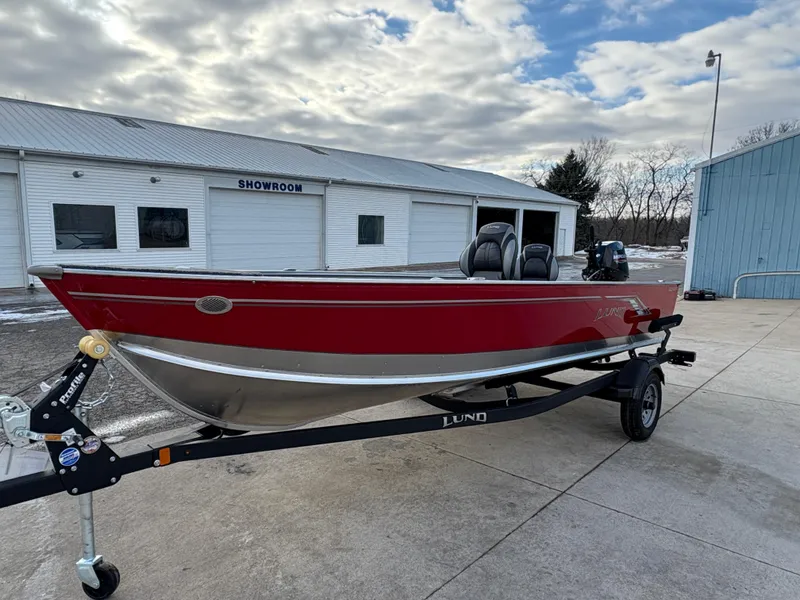 Slide: The Image of 2025 Lund 1600 Fury Tiller boat on trailer outside showroom, under cloudy sky. - 5