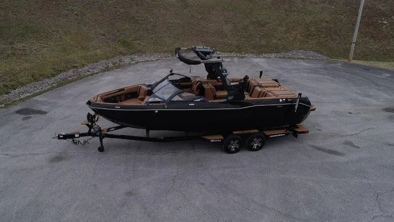 Slide: The Image of 2025 Malibu 25 LSV boat on trailer, black exterior, brown interior, parked on pavement. - 21