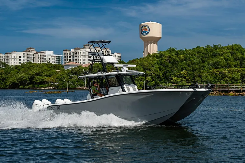 Slide: The Image of 2024 Costa Custom Boats 34 cruising on water near lush greenery and buildings. - 111