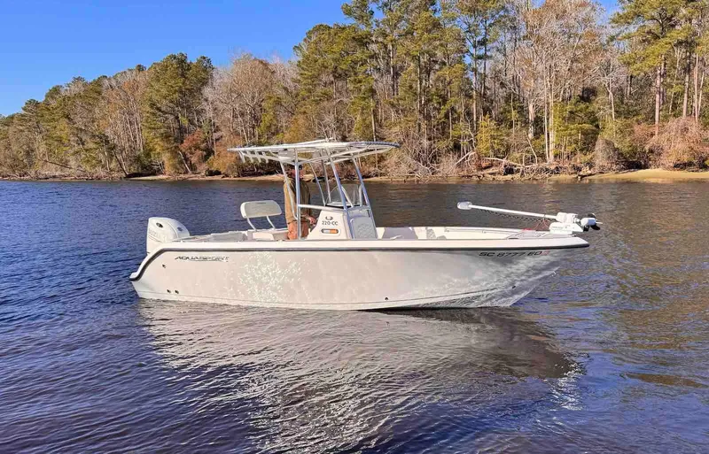 The Image of 2024 Aquasport 2200 Center Console boat on a calm lake with forested shoreline. - 0