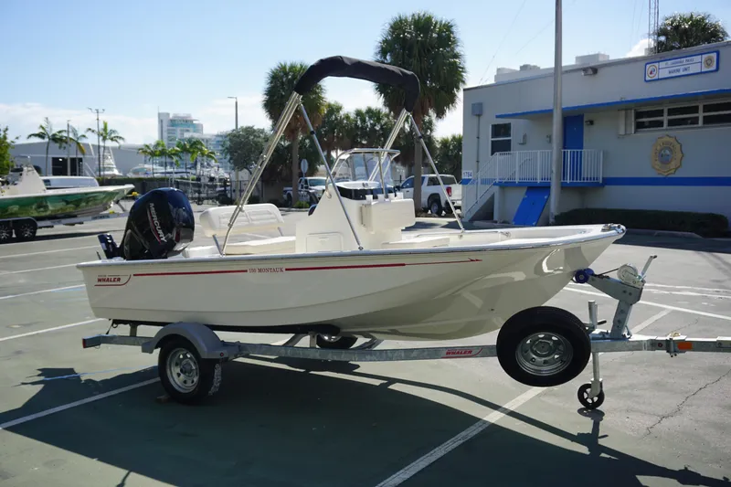 Slide: The Image of 2025 Boston Whaler 150 Montauk boat on trailer, parked outdoors near a building. - 6