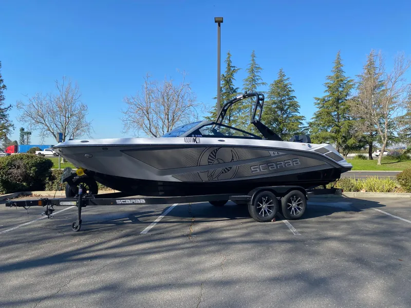 Slide: The Image of 2025 Scarab 235 LX Wake boat on trailer, parked outdoors, clear sky background. - 0