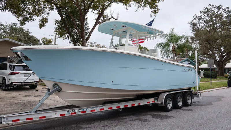 Slide: The Image of 2023 Cobia 280 Center Console boat on a trailer, light blue hull, parked on a street. - 2