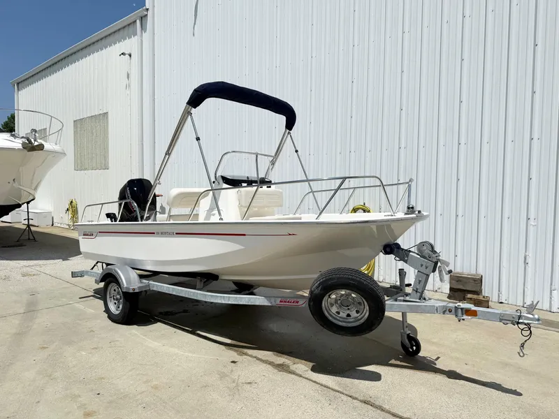 Slide: The Image of 2025 Boston Whaler 150 Montauk boat on trailer, parked near a white building. - 1