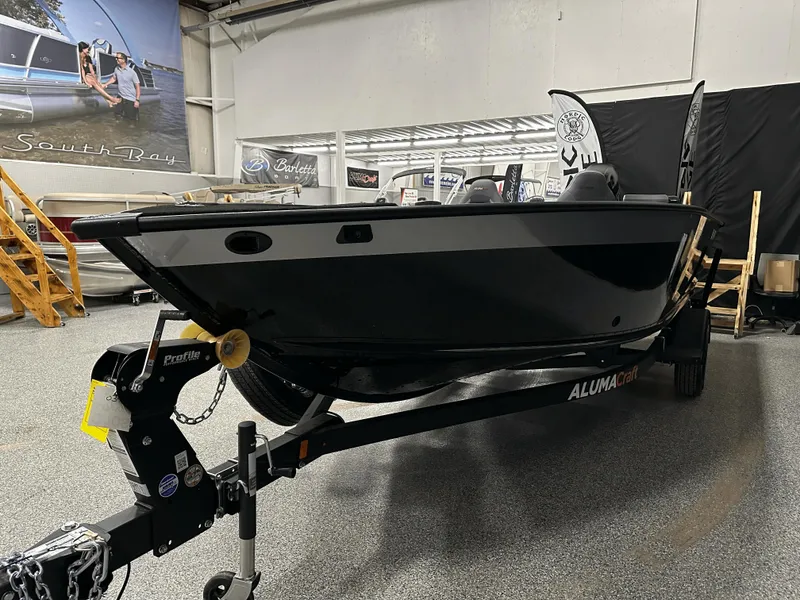 Slide: The Image of 2025 Alumacraft Competitor 185X Tiller boat displayed in showroom, black exterior, on trailer. - 7