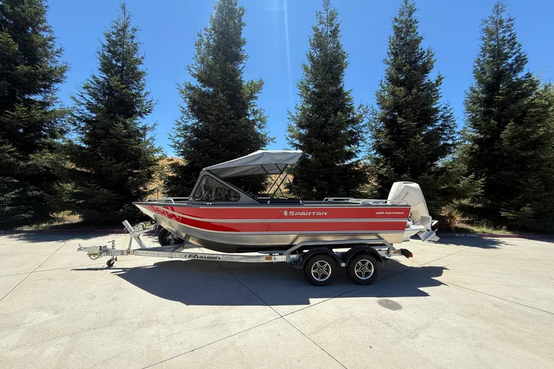 Slide: The Image of 2025 Spartan 200 Astoria boat on trailer, parked outdoors with trees in background. - 1