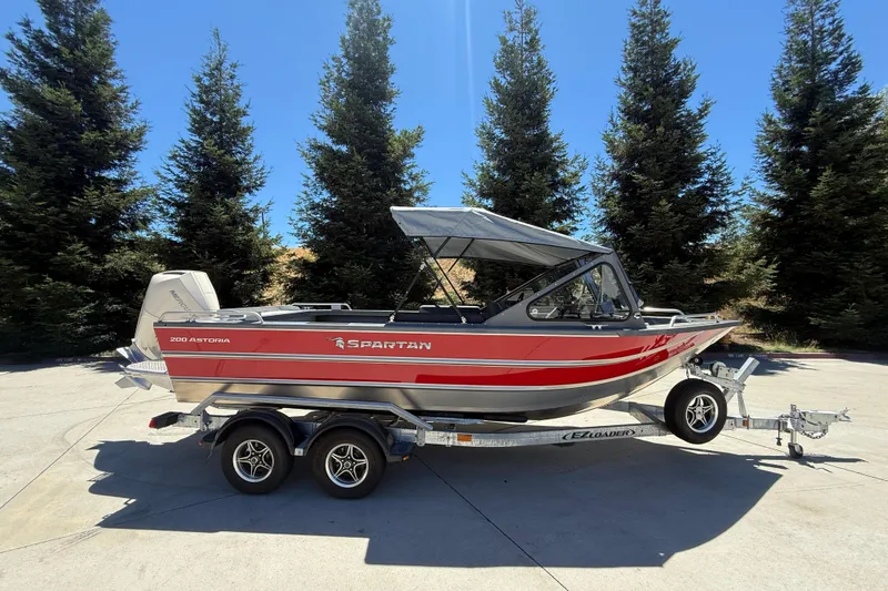 Slide: The Image of 2025 Spartan 200 Astoria boat on trailer, parked outdoors with trees in background. - 0