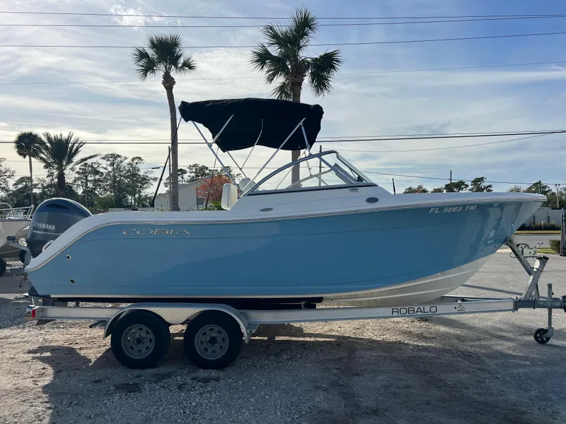 Slide: The Image of 2023 Cobia 220 Dual Console boat on trailer, light blue hull, parked outdoors. - 0