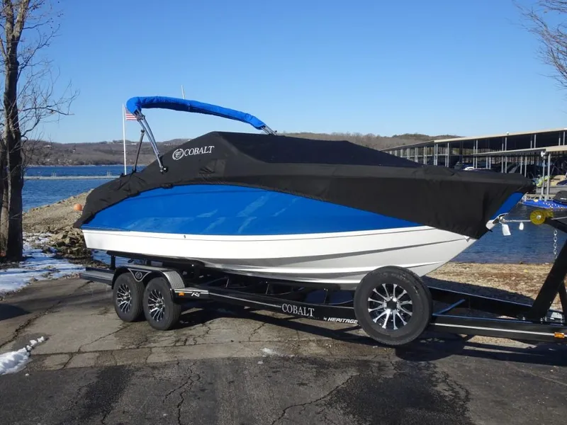 Slide: The Image of 2025 Cobalt R4 boat on trailer by a lake, covered with a black tarp. - 2
