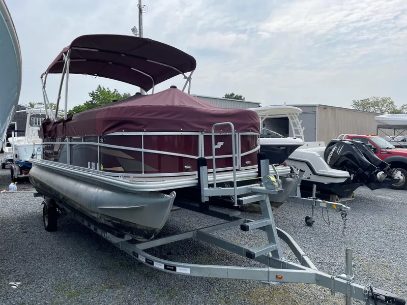 Slide: The Image of 2022 Starcraft LX20R pontoon boat on trailer, maroon canopy, parked in a gravel lot. - 8