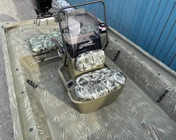 Slide: The Image of 2024 Tracker Grizzly 1860 CC boat with camouflage seats and console, parked near a blue wall. - 9
