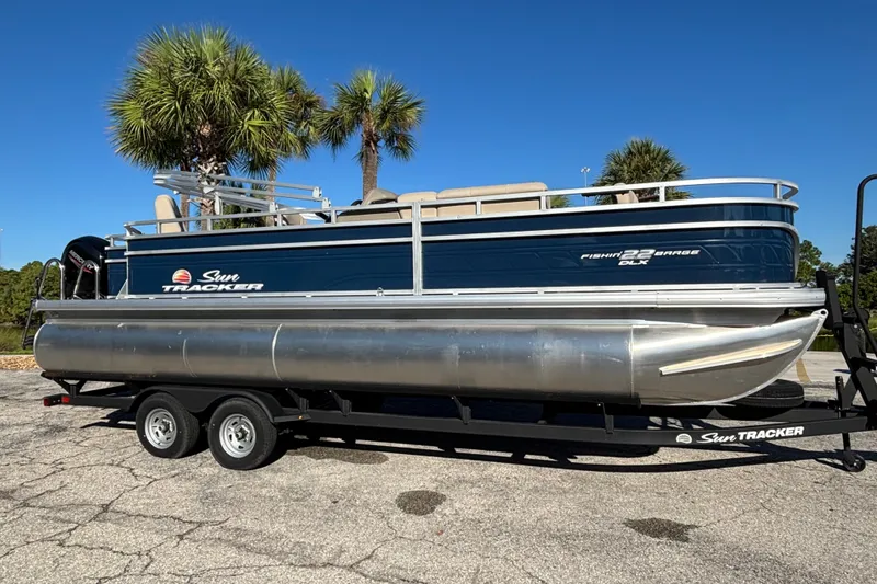 The Image of 2025 Sun Tracker Fishin' Barge 22 DLX pontoon boat on trailer, parked under palm trees. - 0
