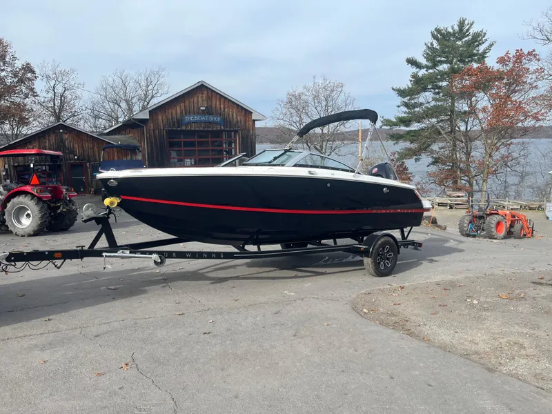 The Image of 2024 Four Winns H1 OB boat on trailer near rustic buildings and trees. - 0