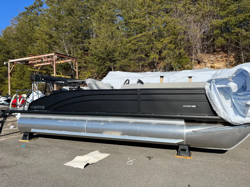 The Image of 2025 Harris Cruiser 230 pontoon boat parked outdoors, partially covered, with trees in the background. - 0