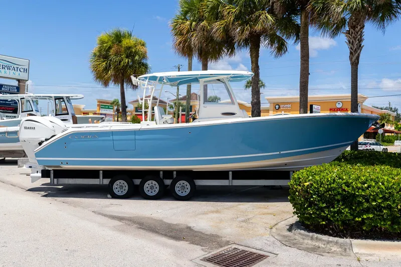 Slide: The Image of 2024 Cobia 320 CC boat on trailer, parked near palm trees and shops. - 1