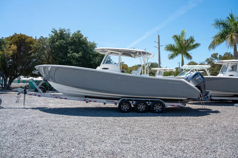 Slide: The Image of 2024 Cobia 320 CC boat on trailer, parked outdoors with trees and clear sky. - 1
