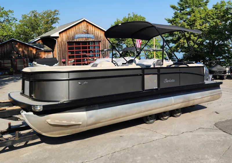 The Image of 2024 Barletta Aria 24QC pontoon boat with canopy, parked outdoors near rustic buildings. - 0