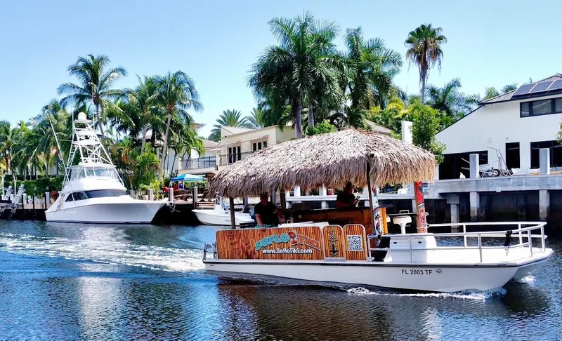 The Image of Beachcat Tiki boat cruising a tropical canal, surrounded by palm trees and waterfront homes. - 1